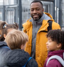friendly teacher welcoming diverse group of students at school gate, backpacks, modern building behind, happy faces, morning light, inclusive education theme